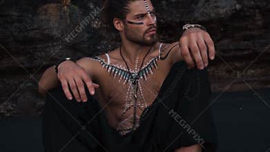 A young, handsome and courageous European man with drawings on his body sits on black sand