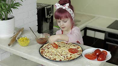 Cooking pizza. Little child in apron adding sliced tomatoes to dough in kitchen