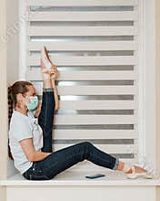 A female ballerina in a mask in casual clothes in jeans and a t-shirt in Pointe shoes at home on the windowsill posing for the