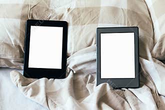 Two modern electronic books with a blank empty screens on a white and beige bed. Mockup tablets on bedding