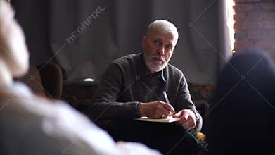 Professional senior man psychologist listening to patient and taking notes on clipboard.
