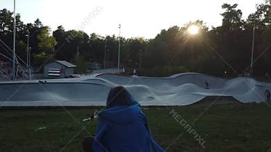 A young woman sitting watching the kids playing with scooters at sunset
