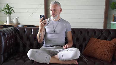 Handsome mature man sitting on the couch at home and communicating with somebody through video chat on his smartphone