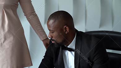 Black man is sitting on chair in white room, dressed in tuxedo, white woman is standing