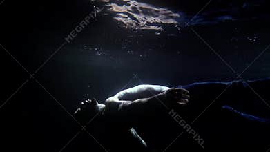 Man is immersing to bottom of pool inside dark water, mysterious light is illuminating his figure