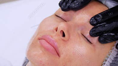 Close-up, cosmetologist, in black medical gloves, performs skincare procedure, facial massage to a woman, using special