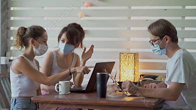 Two young women and man wearing a protective mask sitting at table in cafe with laptop works together