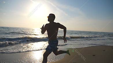 Silhouette of young sporty man running fast along coast during sunrise. Athletic boy training on sea beach. Male