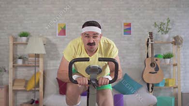 Funny energetic athlete from the 80`s with a mustache engaged at home on a exercise bike slow mo