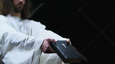 Jesus in robe showing holy bible to camera, faith and belief, dark background