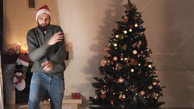 Funny bearded man dancing near the christmas tree. Happy handsome man in Santa`s hat moves his body in funny way. Pretty