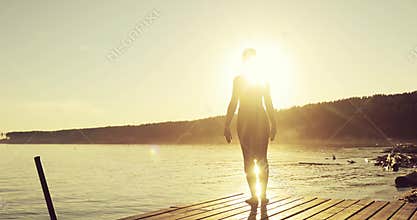 Sporty girl is practicing yoga in tree pose standing on pier by river at sunset.