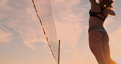 SLOW MOTION, LOW ANGLE, CLOSE UP, SUN FLARE: Athletic girl playing beach volleyball jumps in the air and strikes the