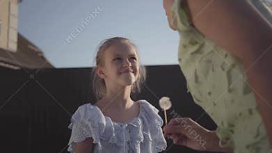 Nice kind daughter giving her mother a dandelion flower and smiling at her in summer evening at village