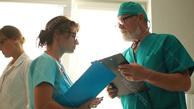 Doctor gives instructions to the assistant. Mature man and young woman in medical gowns talking while standing in a