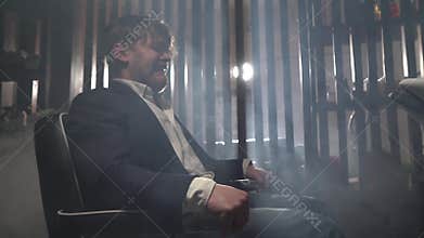 Male businessman in blue jacket and white shirt sits in electric chair and twitches. Torture in gloomy place for young