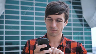 Attractive man standing near big modern office glass building and using smartphone. Red plaid shirt dressed.