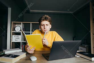 Pensive teenage boy sitting at a table at home with a laptop and books with a notebook and a pen in his hands, looking at camera