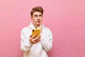 Portrait of thoughtful student boy in white hoodie on pink background holding smartphone, looking away at copy space with pensive