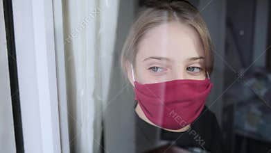 Sad girl wearing a cloth face mask looking through window during worldwide virus epidemic.