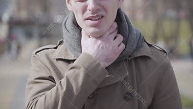 Close-up of young sick man couching and rubbing throat. Portrait of ill Caucasian guy outdoors on sunny autumn or spring