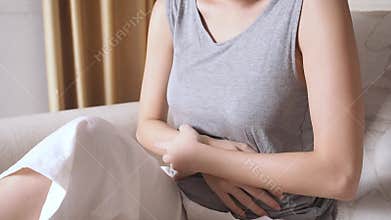 Asian woman sitting on sofa in living room with stomach ache. female belly pain due to pregnancy.