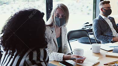 Modern workplace meeting after COVID-19 lockdowns. Young female multiethnic partners talk at office wearing face masks.