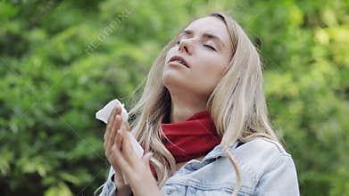 Young woman with with allergy symptom blowing nose standing in the park. Sick girl sneezing and blowing her nose into