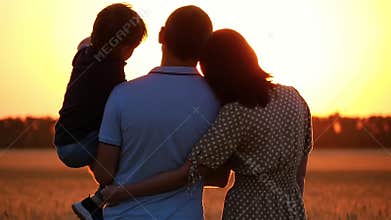 Happy family watching the sunset, standing in a wheat field. A man holding a child in his arms. A woman hugs a man