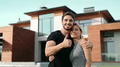 Happy couple making thumbs up near luxury house. Excited family smiling outside