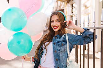 Close-up portrait of gorgeous curly girl in denim jacket posing with birthday balloons outside. Adorable young woman in