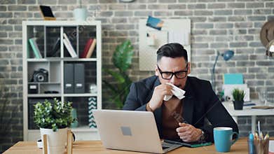 Sick young man using laptop in office then sneezing wiping nose with tissue