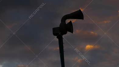 Silhouette of old mechanical civil defense siren, also known as air-raid siren or tornado siren against cloudy sunset sky.