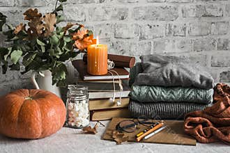 Autumn cozy home still life. Pumpkin, stack of books, burning candle, pile of winter autumn sweaters, jar of marshmallows on the