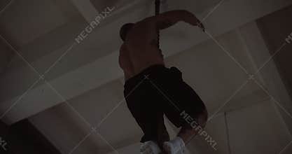 Shirtless young athletic Caucasian sportsman climbing rope during extreme functional physical workout in large gym.