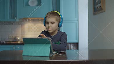 Autistic boy using tablet computer with headphones on the kitchen. Kid with autism looking around at home. Autism