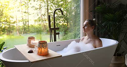 woman relaxing in bathtub full of foam and looking out of window at wellness spa hotel. slider shot