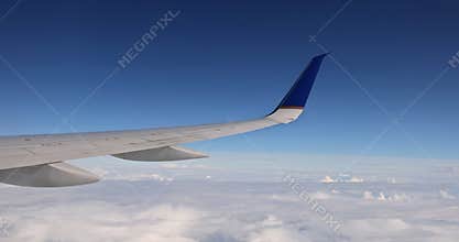 In the background of a fluffy cloudy sky, an airplane wing is seen flying above clouds from a window