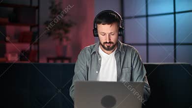 Confident male business freelancer in headphones working at late night use laptop at home interior