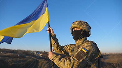 Dolly shot of male soldier in military uniform stands to the peak of hill waving flag of Ukraine. Ukrainian army man