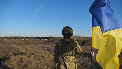 Dolly shot of male soldier in military uniform and helmet holds a waving flag of Ukraine. Ukrainian army man stands with