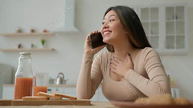 Carefree ethnic Asian woman housewife talking mobile phone in kitchen talk with friend calling speak cellphone share