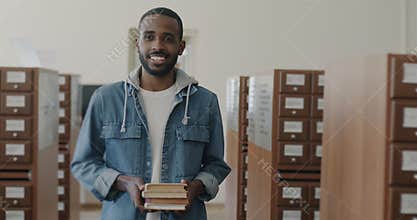 Dolly shot of carefree African American man holding books walking in library hall looking at shelves
