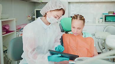 Woman dentist shows educational video on tablet to teen girl patient.