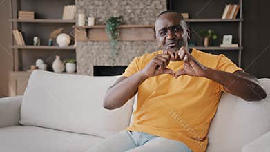 Mature kind gentle african american man rest time at home living room put folded palms on heart looking at camera shows