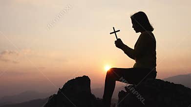 A silhouette of a woman with a cross in her hands praying to God. while the sun was rising. Praise the religion. highest faith in