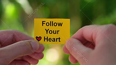 A woman holding a yellow paper note with the phrase Follow Your Heart written on it in her hands against a foliage background.
