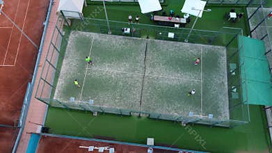 Aerial drone view of a padel tennis match in progress, surrounded by red clay tennis courts and green walkways