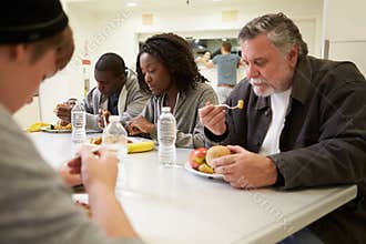 People Sitting At Table Eating Food In Homeless Shelter
