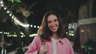 A joyful, smiling woman gracefully walks through a vibrant outdoor setting at night, fully enjoying the charming